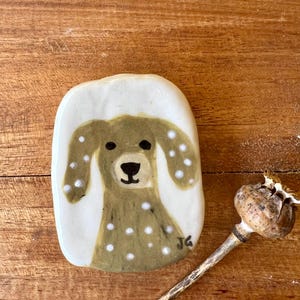May include: A ceramic dog-shaped ornament with a white background and a brown dog design with white spots. The dog has black eyes and a black nose. The ornament is on a wooden surface. A dried seed pod is next to the ornament.