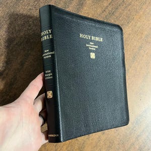 May include: Black leather-bound Holy Bible with gold lettering. The spine reads "Holy Bible" and "New International Version, Wide Margin Edition." The cover also features gold text. The book is held open.