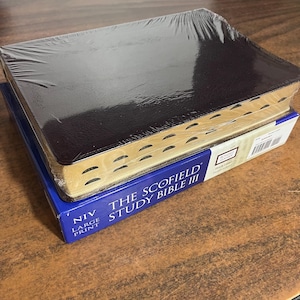 May include: A new, sealed black leather-bound Bible with gold-gilded edges, resting on a blue box. The box reads "The Scofield Study Bible III" and "NIV Large Print". The Bible is wrapped in clear plastic.