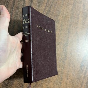 May include: A burgundy leather-bound Holy Bible with gold lettering. The spine reads "Holy Bible" and "New International Version." The Zondervan logo is visible at the bottom of the spine. A hand is holding the book.