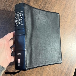 May include: A black NIV Study Bible by Zondervan. The book has a flexible cover and a black ribbon bookmark. The spine features the text "NIV STUDY BIBLE" in white. The book is held by a person.