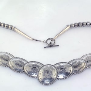 May include: A silver-toned necklace featuring a series of oval pendants with etched floral designs. The necklace has a beaded chain and a toggle clasp. The design is symmetrical and the metal has a polished finish.