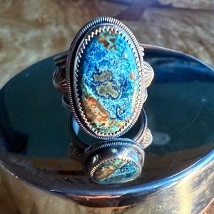 May include: A silver ring with a large oval turquoise stone set in a silver bezel. The stone has a blue, green, and brown pattern.