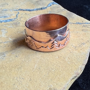 May include: A wide copper ring featuring a polished finish and a textured band with an embossed landscape design depicting mountain peaks and evergreen trees. The metal has a warm, reddish-brown metallic luster and a smooth, rounded interior surface.