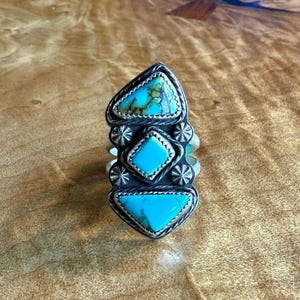 May include: A silver ring with three turquoise stones set in a geometric design. The stones are a light blue color and are surrounded by silver.