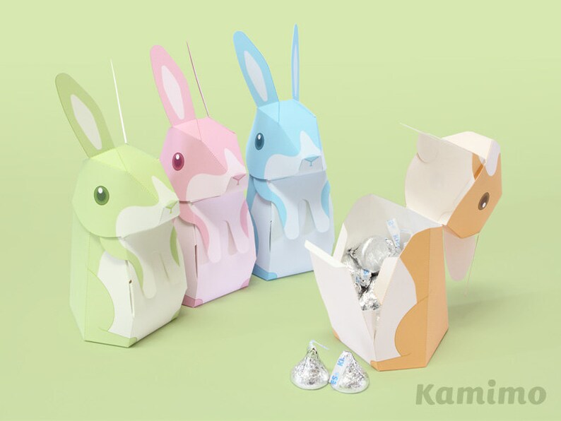 Puede incluir: Cuatro cajas de papel con forma de conejo, una abierta para revelar caramelos de chocolate en su interior. Las cajas son verdes, rosas, azules y naranjas. Las cajas est&aacute;n etiquetadas con "Kamimo".