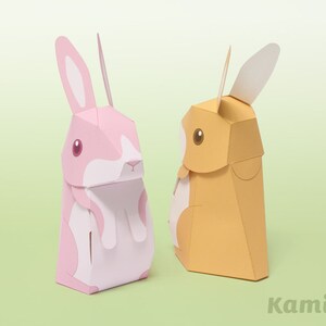 Puede incluir: Dos figuritas de conejo de papel, una rosa y otra naranja, de pie sobre un fondo verde. El conejo rosa tiene orejas blancas y una cara blanca con detalles rosas. El conejo naranja tiene orejas blancas y una cara blanca con detalles marrones. La palabra "Kamimo" est&aacute; impresa en la esquina inferior derecha de la imagen.