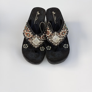 May include: A pair of black flip-flops with a floral design and rhinestone embellishments. The flip-flops have a wedge heel and say "Montana West" on the straps.