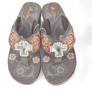 May include: A pair of brown flip-flops with orange and silver embroidery. The flip-flops have a large rhinestone cross on the strap and the words "Montana West" on the sole.