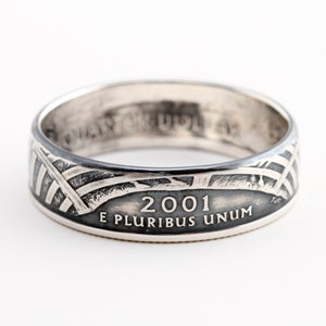 May include: A silver ring with a design featuring the year 2001 and the inscription "E PLURIBUS UNUM." The ring has a band with a pattern of intersecting lines and a flag design. The ring is likely made from a quarter coin.