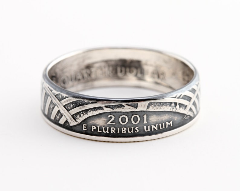 May include: A silver ring with a design featuring the year 2001 and the inscription "E PLURIBUS UNUM." The ring has a band with a pattern of intersecting lines and a flag design. The ring is likely made from a quarter coin.