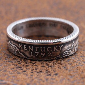 May include: A silver ring with the word "Kentucky" and the year "1792" engraved on it. The ring has a detailed design and a textured surface. The ring is a piece of jewelry.