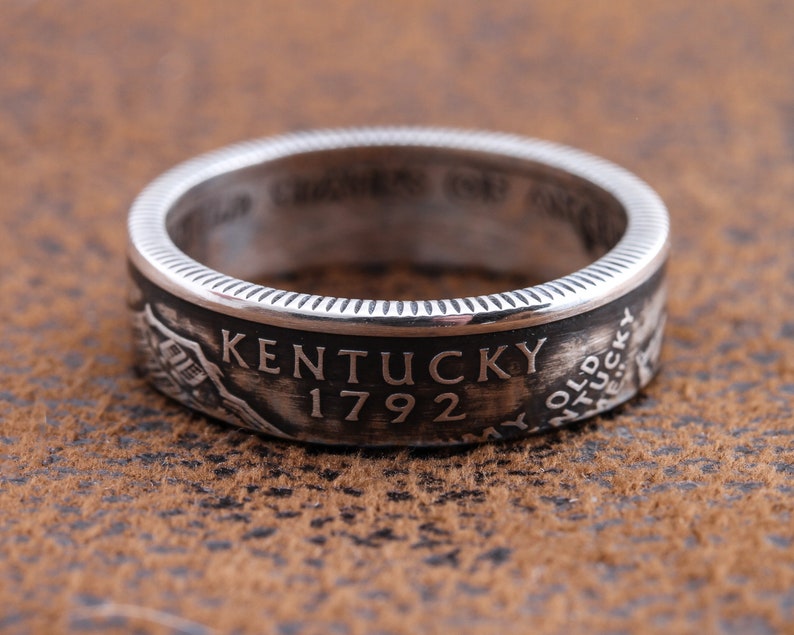 May include: A silver ring with the word "Kentucky" and the year "1792" engraved on it. The ring has a detailed design and a textured surface. The ring is a piece of jewelry.