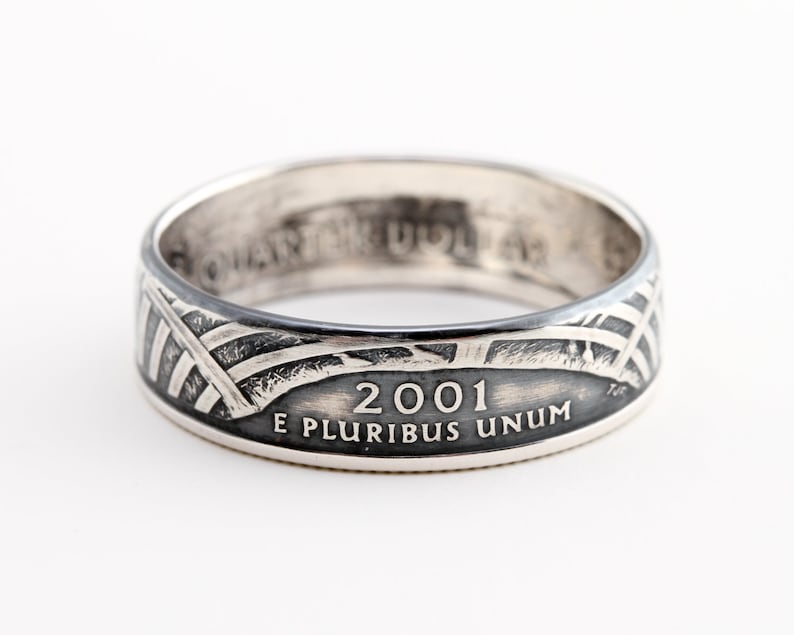 May include: A silver ring with a design featuring the year 2001 and the inscription "E PLURIBUS UNUM." The ring has a band with a pattern of intersecting lines and a flag design. The ring is likely made from a quarter coin.