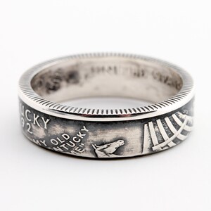May include: A silver ring with a detailed design. The ring features the text "Kentucky 1992" and a horse head. The ring has a textured edge and a polished finish. The ring is a piece of jewelry.