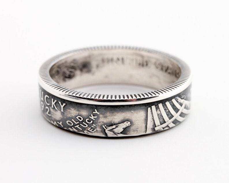 May include: A silver ring with a detailed design. The ring features the text "Kentucky 1992" and a horse head. The ring has a textured edge and a polished finish. The ring is a piece of jewelry.