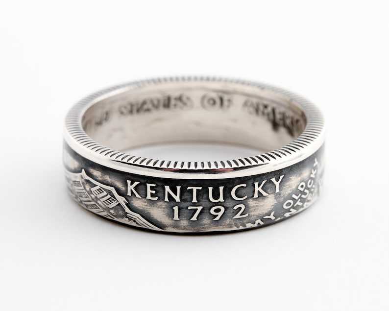 May include: A silver ring with the text "KENTUCKY 1792" and an image of a building. The ring has a detailed, textured design around the edges and is set against a white background.