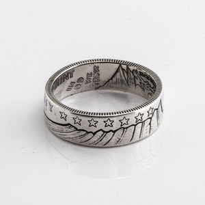 May include: A silver ring with engraved details. The ring features a mountain range design with stars and the text "IN GOD WE TRUST" and "LIBERTY". The ring is likely made from a coin.