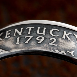 May include: Close-up of a silver ring with the word "KENTUCKY" and the number "1792" engraved on it. The ring has a dark, textured background and a polished silver finish. The ring is likely a commemorative or historical piece.
