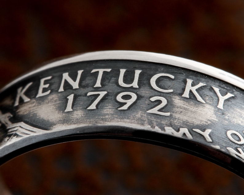May include: Close-up of a silver ring with the word "KENTUCKY" and the number "1792" engraved on it. The ring has a dark, textured background and a polished silver finish. The ring is likely a commemorative or historical piece.