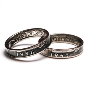May include: Two silver rings made from quarter dollars, with the dates 1996 and 1965 visible. The rings are set against a white background, showcasing the intricate details of the coin's design and the text "QUARTER DOLLAR".