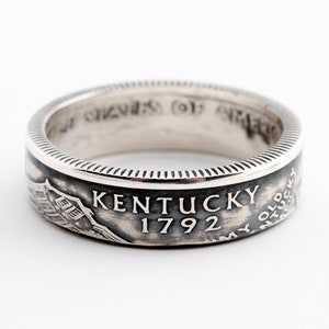 May include: A silver ring with the text "KENTUCKY 1792" and an image of a building. The ring has a detailed, textured design around the edges and a dark background for the text and image.