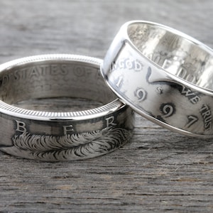 May include: Two silver rings made from coins, resting on a wooden surface. The rings display embossed text and designs, including the words "LIBERTY" and "IN GOD WE TRUST". The rings have a diameter of approximately 1 inch.