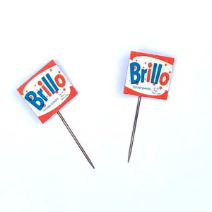 May include: Two red and white Brillo soap pads with blue lettering on a white background. The soap pads are attached to small metal pins.