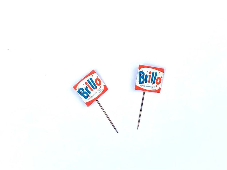 May include: Two red and white Brillo soap pads with blue lettering on a white background. The soap pads are attached to small metal pins.