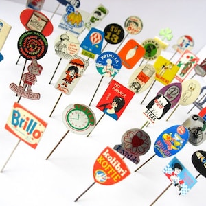 May include: A collection of vintage advertising pins featuring various brands and logos. The pins are colorful and feature a variety of designs, including cartoon characters, text, and images. Some of the brands featured include "Brillo", "Pastachoca", "Hit Parade", "Primula", "Green Spot", and "Popla".