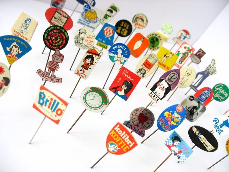 May include: A collection of vintage advertising pins featuring various brands and logos. The pins are colorful and feature a variety of designs, including cartoon characters, text, and images. Some of the brands featured include "Brillo", "Pastachoca", "Hit Parade", "Primula", "Green Spot", and "Popla".