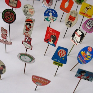 May include: A collection of vintage advertising pins featuring various brands and products. The pins are colorful and feature illustrations of people, animals, and objects. Some of the brands featured include 7-Up, Wella, King Rexi, and Diddas Dekens.