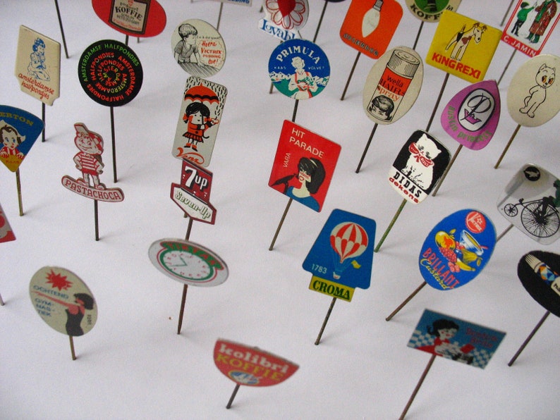 May include: A collection of vintage advertising pins featuring various brands and products. The pins are colorful and feature illustrations of people, animals, and objects. Some of the brands featured include 7-Up, Wella, King Rexi, and Diddas Dekens.