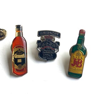 May include: Three metal bottle opener pins featuring the logos for Smirnoff Vodka, Grants 12, and J&B Rare Scotch Whisky. The pins are on a white background.
