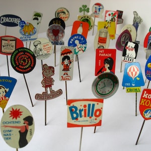 May include: A collection of vintage advertising pins featuring various brands and products, including "Karpeton", "Brillo", "Kolibri Koffie", "Pastachoca", "Croma", "Didias", "Hit Parade", "Primula", "Sus", "Crackfree", "Ochtend Gym Nas Tiek", and "Brilliant Custard".