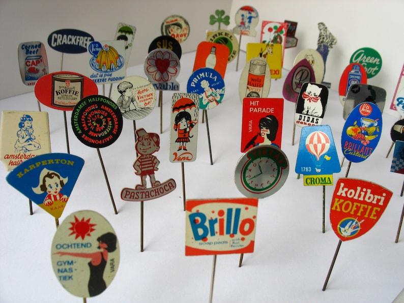 May include: A collection of vintage advertising pins featuring various brands and products, including "Karpeton", "Brillo", "Kolibri Koffie", "Pastachoca", "Croma", "Didias", "Hit Parade", "Primula", "Sus", "Crackfree", "Ochtend Gym Nas Tiek", and "Brilliant Custard".