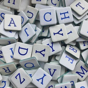 Può includere: Primo piano di un mucchio di tessere bianche di Scrabble con lettere blu. Le tessere sono sparse, mostrando varie lettere come A, C, Y e Z. L'immagine è ben illuminata, evidenziando il design classico dei pezzi del gioco.