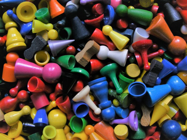 May include: A close-up shot of a collection of colorful game pieces. The pieces are various shapes and sizes, including pawns, cones, and cylinders, in shades of red, yellow, green, blue, pink, and orange. Some are wooden, others plastic.