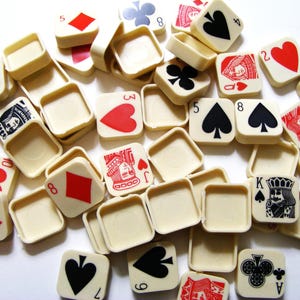 Pode incluir: Uma cole&ccedil;&atilde;o de pe&ccedil;as quadradas de cartas de jogar em branco sujo com s&iacute;mbolos de paus, ouros, copas e espadas vermelhos, pretos e verdes. As pe&ccedil;as apresentam n&uacute;meros e figuras, incluindo o &Aacute;s, o Rei, a Dama e o Valete. Espalhadas.