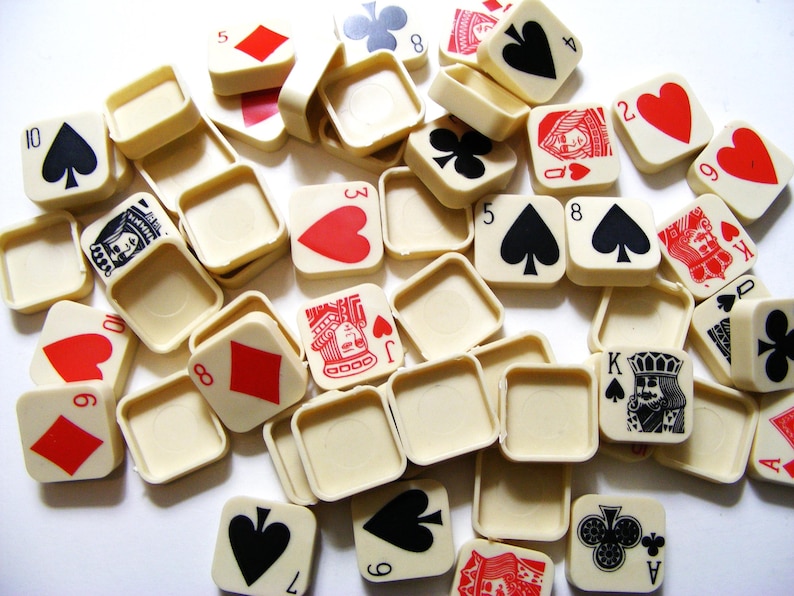 Pode incluir: Uma cole&ccedil;&atilde;o de pe&ccedil;as quadradas de cartas de jogar em branco sujo com s&iacute;mbolos de paus, ouros, copas e espadas vermelhos, pretos e verdes. As pe&ccedil;as apresentam n&uacute;meros e figuras, incluindo o &Aacute;s, o Rei, a Dama e o Valete. Espalhadas.