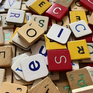 May include: A collection of square wooden game tiles scattered in a pile. The tiles feature various letters of the alphabet printed in black, blue, red, or green, with small numerical values in the corners of some tiles, showing a mix of colours.