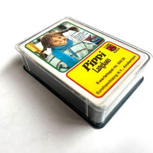 Op de afbeelding: Een kaartspel van Pippi Langkous in een doorzichtige plastic hoes. De bovenste kaart toont een portret van een jong meisje met vlechtjes, met een blauwe sjaal en een trui met patroon. Het gele label van de kaart luidt "Pippi Langkous".