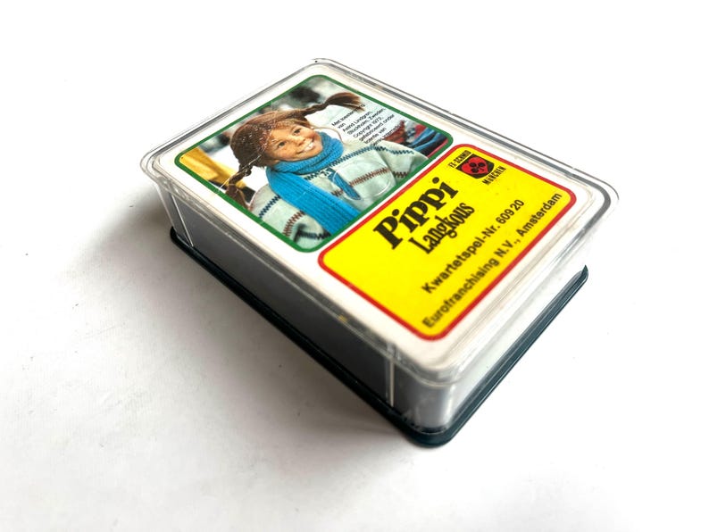 Op de afbeelding: Een kaartspel van Pippi Langkous in een doorzichtige plastic hoes. De bovenste kaart toont een portret van een jong meisje met vlechtjes, met een blauwe sjaal en een trui met patroon. Het gele label van de kaart luidt "Pippi Langkous".
