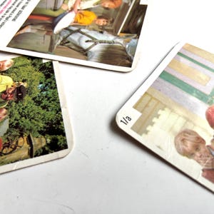 Op de afbeelding: Drie vintage speelkaarten met afgeronde hoeken liggen verspreid op een wit oppervlak. Elke kaart heeft een kleurrijke afbeelding van mensen. E&eacute;n kaart heeft het nummer "1/a" in de hoek. De kaarten lijken uit een kinderspel te komen.