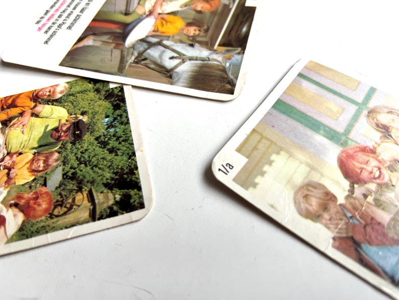 Op de afbeelding: Drie vintage speelkaarten met afgeronde hoeken liggen verspreid op een wit oppervlak. Elke kaart heeft een kleurrijke afbeelding van mensen. E&eacute;n kaart heeft het nummer "1/a" in de hoek. De kaarten lijken uit een kinderspel te komen.