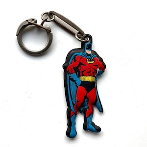 Puede incluir: Un llavero de goma rojo y azul con una imagen de dibujos animados de Batman. El llavero tiene un anillo de metal plateado y un cierre de metal plateado.