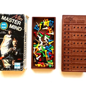 May include: A Mini Master Mind board game set including a rectangular box with a photo of two people, a brown plastic tray filled with assorted colorful plastic pegs, and a brown plastic game board with numbered rows and holes for the pegs.