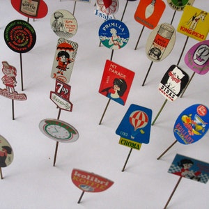 Puede incluir: Una colecci&oacute;n de pines publicitarios vintage con varios dise&ntilde;os y colores. Los pines presentan im&aacute;genes de productos, marcas y lemas. Algunos de los pines incluyen texto como "7up", "Hit Parade", "King Rexi", "Didas", "Brilliant", "Kolibri Koffie" y "1783 Croma".
