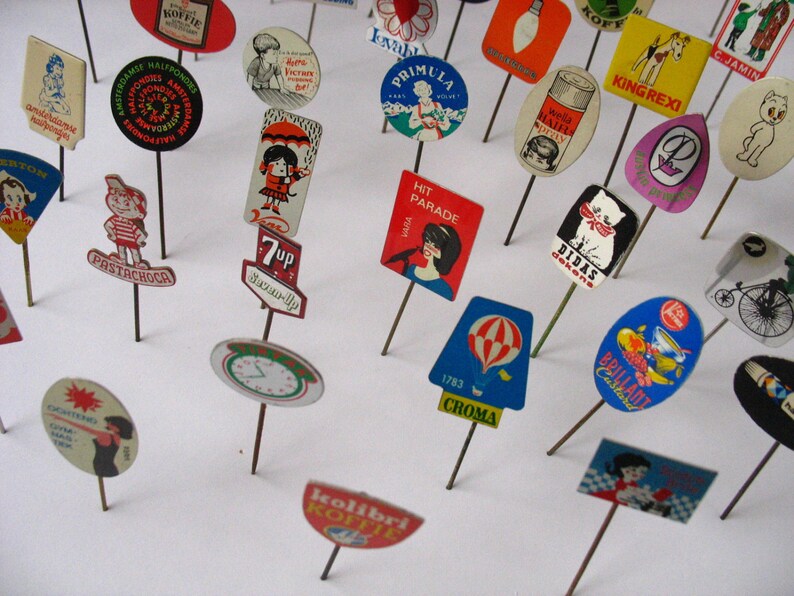 Puede incluir: Una colecci&oacute;n de pines publicitarios vintage con varios dise&ntilde;os y colores. Los pines presentan im&aacute;genes de productos, marcas y lemas. Algunos de los pines incluyen texto como "7up", "Hit Parade", "King Rexi", "Didas", "Brilliant", "Kolibri Koffie" y "1783 Croma".