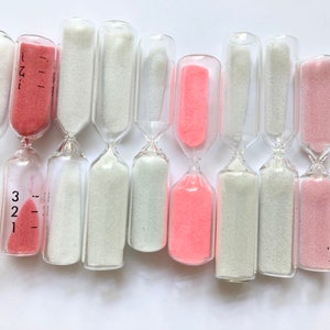 Könnte beinhalten: Eine Reihe von 12 Sanduhren, einige mit rosa Sand gefüllt, andere mit weißem Sand. Die Sanduhren sind alle aus klarem Glas und haben einen schmalen Hals in der Mitte.